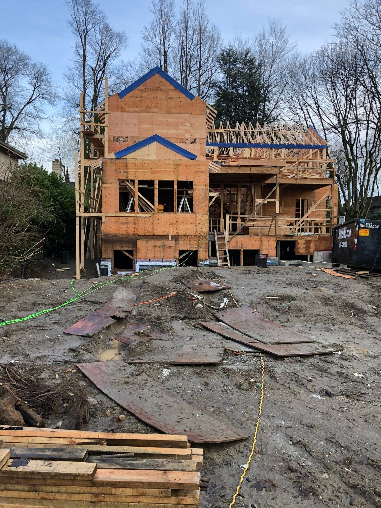 Partially constructed house with wooden framing, located on a muddy plot with construction materials scattered around.